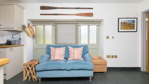 A room with a blue sofa, a wooden table, a window with blinds, a framed picture on the wall, and a door. - Scilly Stack (Penzance)