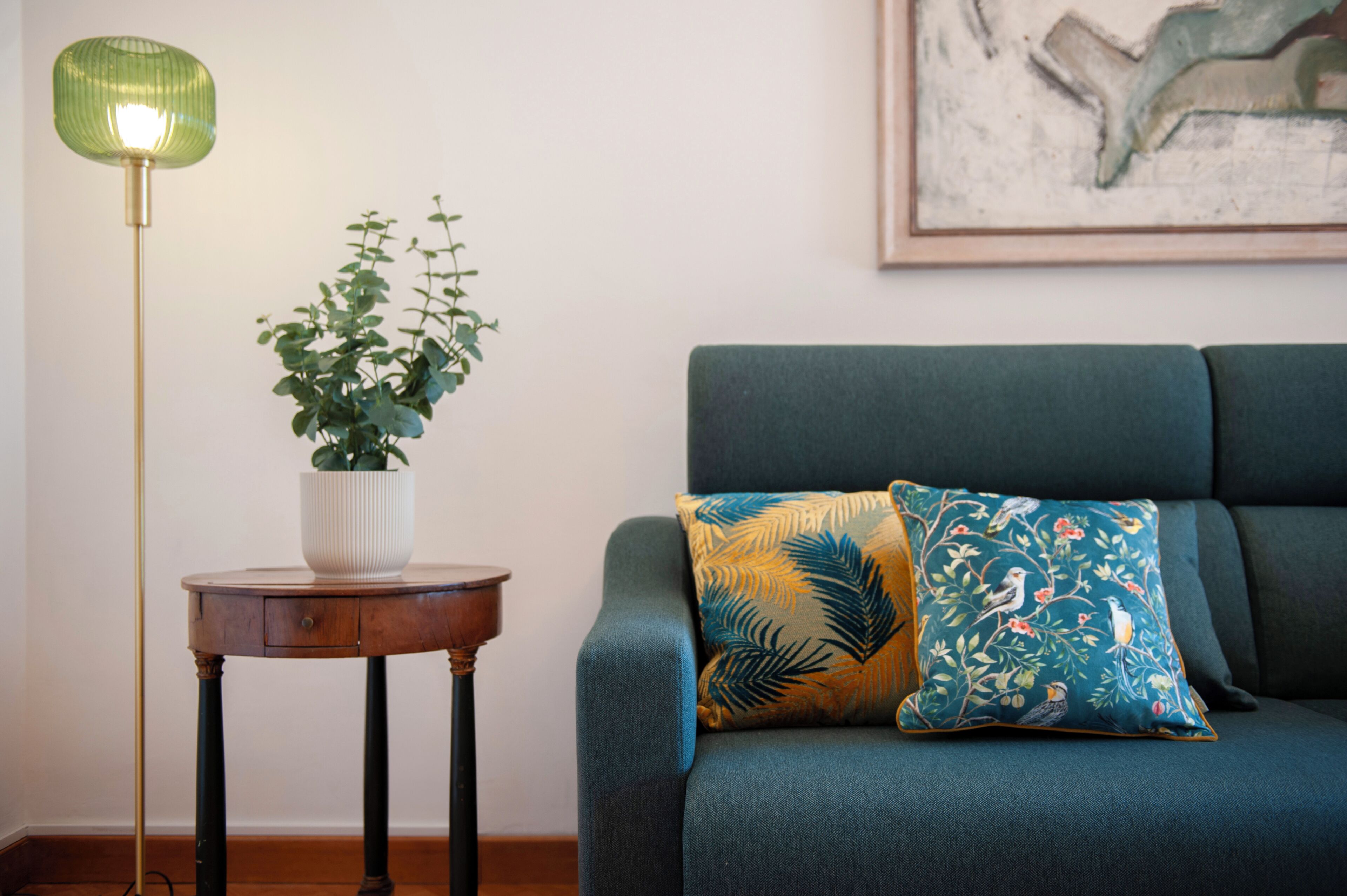 A room with a green sofa, a small round wooden table with a white vase and green plant, and a green floor lamp.