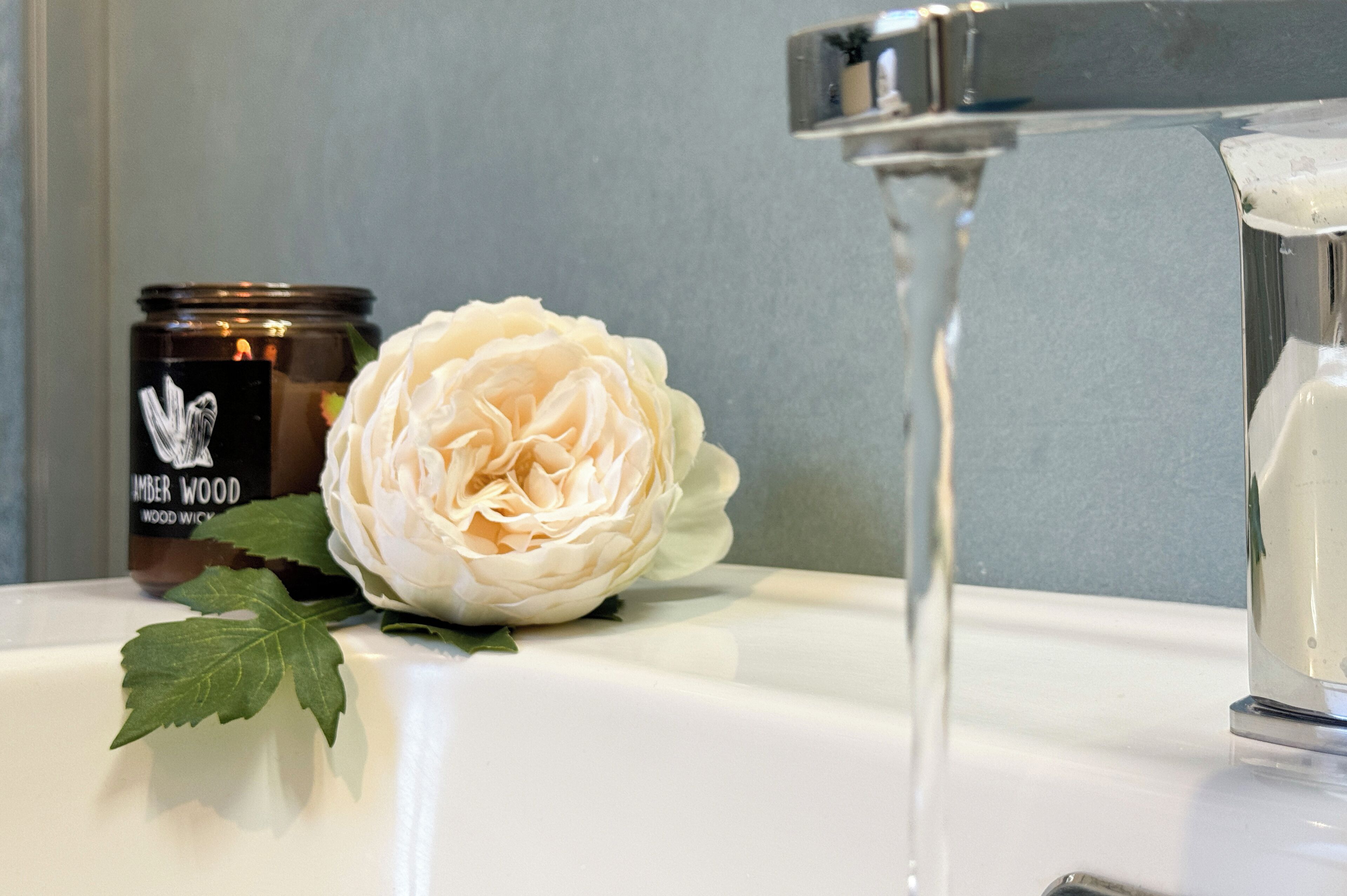 A bathroom sink with a white rose, a jar labeled 'LUMBER WOOD,' and a clear glass.