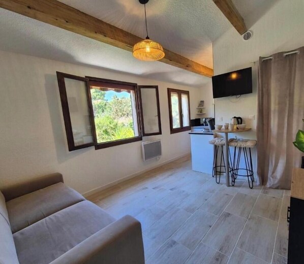 A room with a sofa, a dining area with stools, a TV, and windows with a view of greenery. - 🏖️ Studio swimming pool 300 meters from the sea 🌊 (Six-Fours-les-Plages)