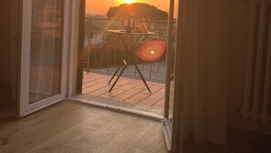 Puerta corredera abierta que da a un balcón con vistas a la puesta de sol y una mesa con sillas.