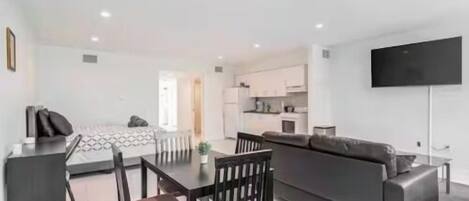 Una sala de estar moderna con sofá, mesa de comedor y sillas. Hay una zona de cocina con armarios y un televisor de pantalla plana montado en la pared.