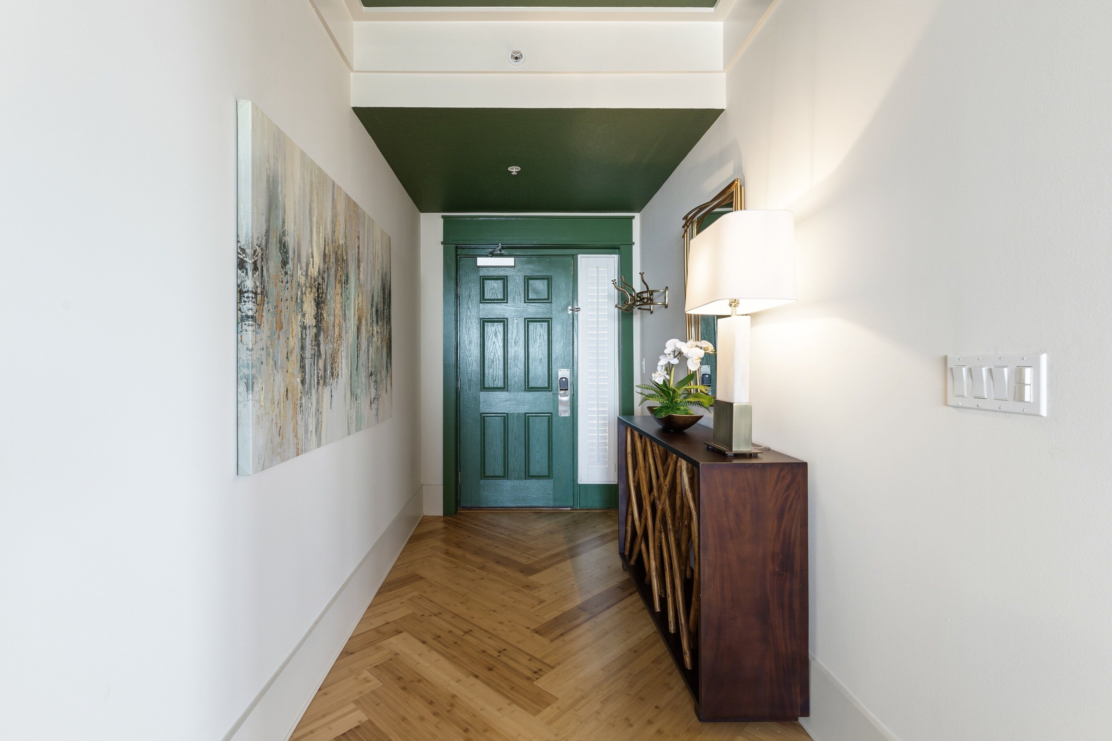 A hallway with a wooden floor, a green door, a wall-mounted lamp, and a painting.