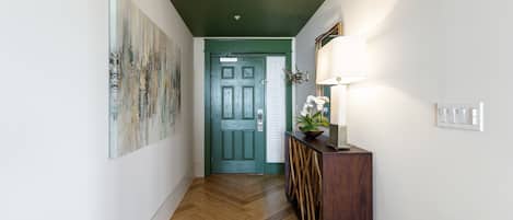 A hallway with a wooden floor, a green door, a wall-mounted lamp, and a painting.