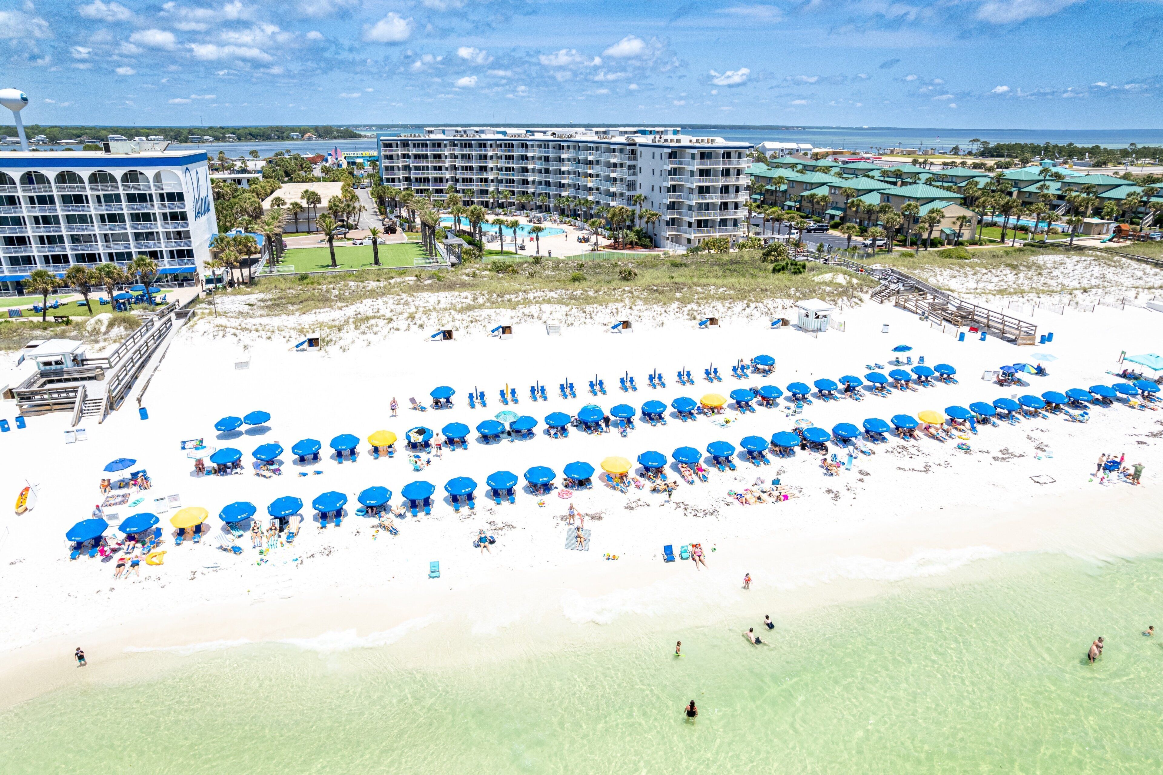 A beach resort with multiple hotel buildings, a sandy beach with numerous umbrellas, and a clear view of the ocean.