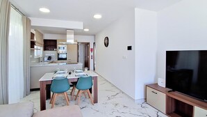 A modern living room with a flat-screen TV, a dining area with two chairs, and a kitchen with a microwave and oven. - Modern Penthouse Retreat with Rooftop City Views (Il-Mellieħa)