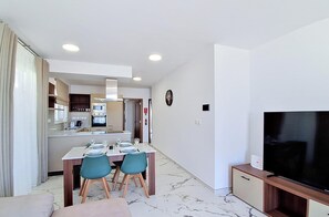 A modern living room with a flat-screen TV, a dining area with a table set for four, and a kitchen with built-in appliances. - Modern Penthouse Retreat with Rooftop City Views (Il-Mellieħa)