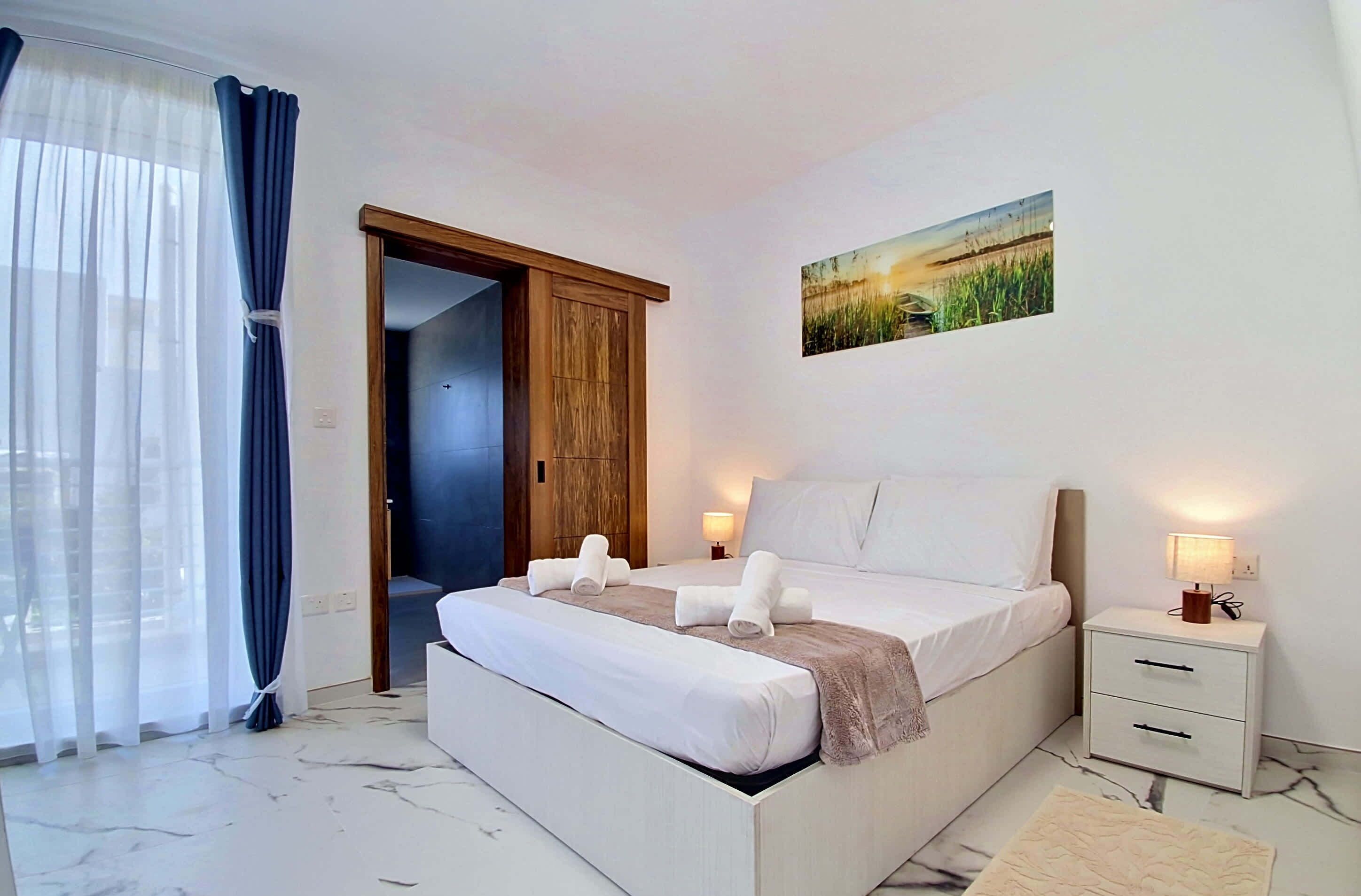 Um quarto com cama, mesas de cabeceira, um criado-mudo, uma porta e uma janela com cortinas.