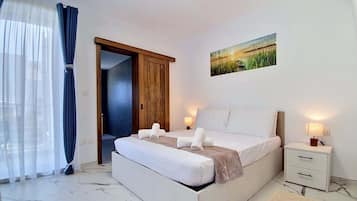 Um quarto com cama, mesas de cabeceira, um criado-mudo, uma porta e uma janela com cortinas.