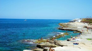 A coastal beach with rocky formations, clear blue water, and a distant sailboat. - Bright Sliema Coastal Apartment with Sea Terrace (Tas-Sliema)