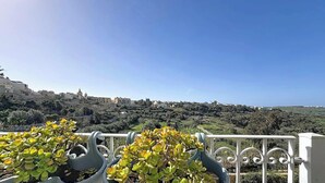 A balcony with potted plants, a clear sky, and a distant view of a town with buildings and greenery. - Scenic Gozo Apartment & Shared Pool (Ix-Xagħra)