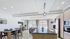 A modern kitchen with a central island, a dining area with a table and chairs, and a living space with a sofa and large windows. - Scenic Gozo Apartment & Shared Pool (Ix-Xagħra)