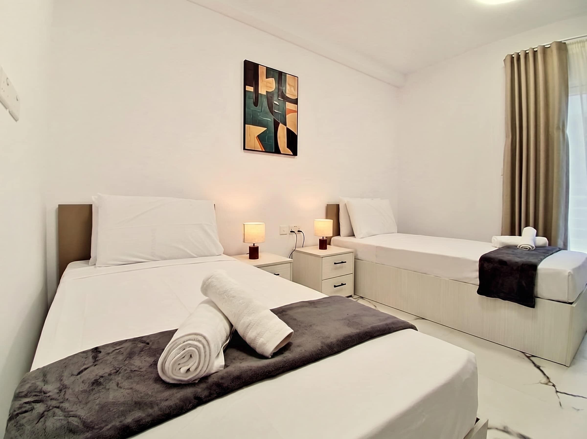 A hotel room with two beds, a nightstand with lamps, a painting on the wall, and a window with curtains.
