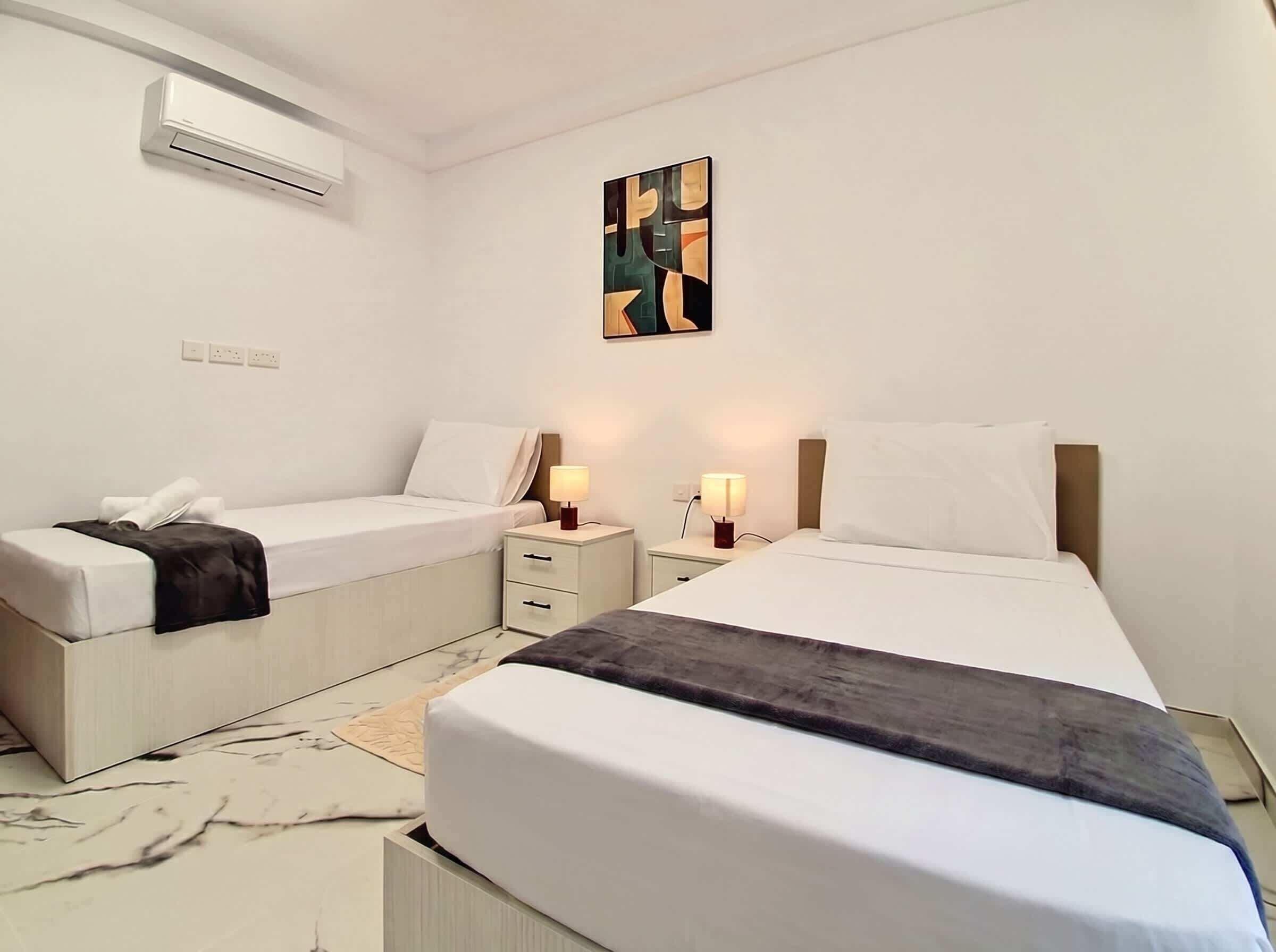 A hotel room with two beds, a bedside table with lamps, a wall-mounted air conditioner, and a painting.