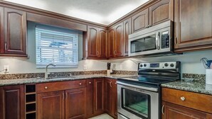 A kitchen with wooden cabinets, a microwave, an oven, granite countertops, and a window with blinds.