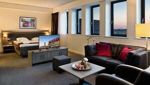 Ein Hotelzimmer mit Bett, Fernseher, Sofa, Couchtisch und Blick auf die Stadt durch große Fenster.