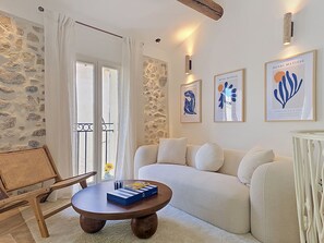 A living room with a sofa, a round wooden coffee table, a wicker chair, and a stone wall. There are framed artworks on the wall and a window with curtains. - New -Vieil Antibes Remparts -Rare House 3CHR/3SDB (Antibes)