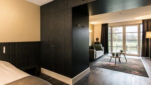 A hotel room with a dark wood wardrobe, a bed, a sofa, a small table with a lamp, and a view through a large window. - Escape to Nature | Scenic Hotel Room with Views of the Dutch Forest! (Berg en Dal)
