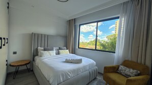 A modern bedroom with a large bed, a small round table, a sofa, and a window with a view of buildings and trees. - Welcoming 2-bedroom apartment with fitness room in Nairobi (Nairobi)