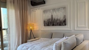 Moderni hotellihuone, jossa on suuri sänky, yöpöytä, lamppu ja kaupunkimaisemaa esittävä taulu seinällä.