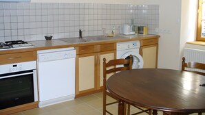 A kitchen with a stove, dishwasher, and washing machine, wooden cabinets, and a round dining table. - “Gîte Les Bannes” with Mountain View and Wi-Fi (Saint-Sauves-d'Auvergne)