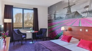 Habitación de hotel con cama, dos sillas, una mesita, lámpara y ventana grande.