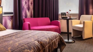 Hotellihuone, jossa on sänky, vaaleanpunainen sohva, keltainen nojatuoli, pieni pöytä, jossa on radio, ja ikkuna, jossa on verhot.