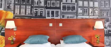 A hotel room with a wooden headboard, two beds with white linens, teal pillows, and a nightstand with a lamp and vase of flowers.