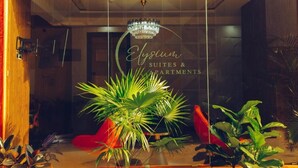 Entrada de um hotel com uma placa indicando 'Elysium Suites & Apartments', um lustre e plantas em vasos.