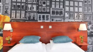 Une chambre d’hôtel avec une tête de lit en bois, deux lits avec du linge de lit blanc, des oreillers turquoise et une table de chevet avec une lampe et un vase de fleurs.