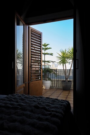 Een kamer met een bed, een balkon met uitzicht op palmbomen en een hek, en een grote kamerplant.