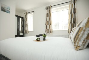 Ein Schlafzimmer mit einem großen Bett, einem Nachttisch mit Pflanze, einem kleinen Tisch mit Tasse und Buch sowie einem Fenster mit Vorhängen.