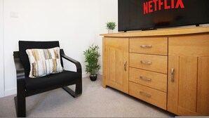 Ett vardagsrum med en svart soffa, ett träskåp, en tv-apparat som visar "NETFLIX" och en liten krukväxt.