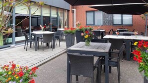 Une terrasse extérieure avec des tables et des chaises, des plantes en pot et un bâtiment en arrière-plan.