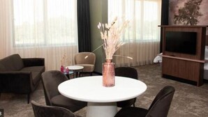 Une chambre d’hôtel comprenant une table à manger ronde, deux canapés, un meuble pour la télévision et un vase contenant des fleurs séchées.