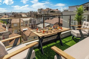 Eine Dachterrasse mit Esstisch, gedecktem Tisch für eine Mahlzeit, umgeben von Sitzgelegenheiten mit Kissen und Blick auf eine Stadtlandschaft.