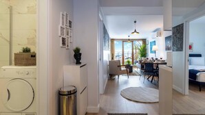 Moderni hotellihuone, jossa on pyykinpesukone, roskakori, ruokailutila pöytineen ja tuoleineen sekä näkymä ulos.
