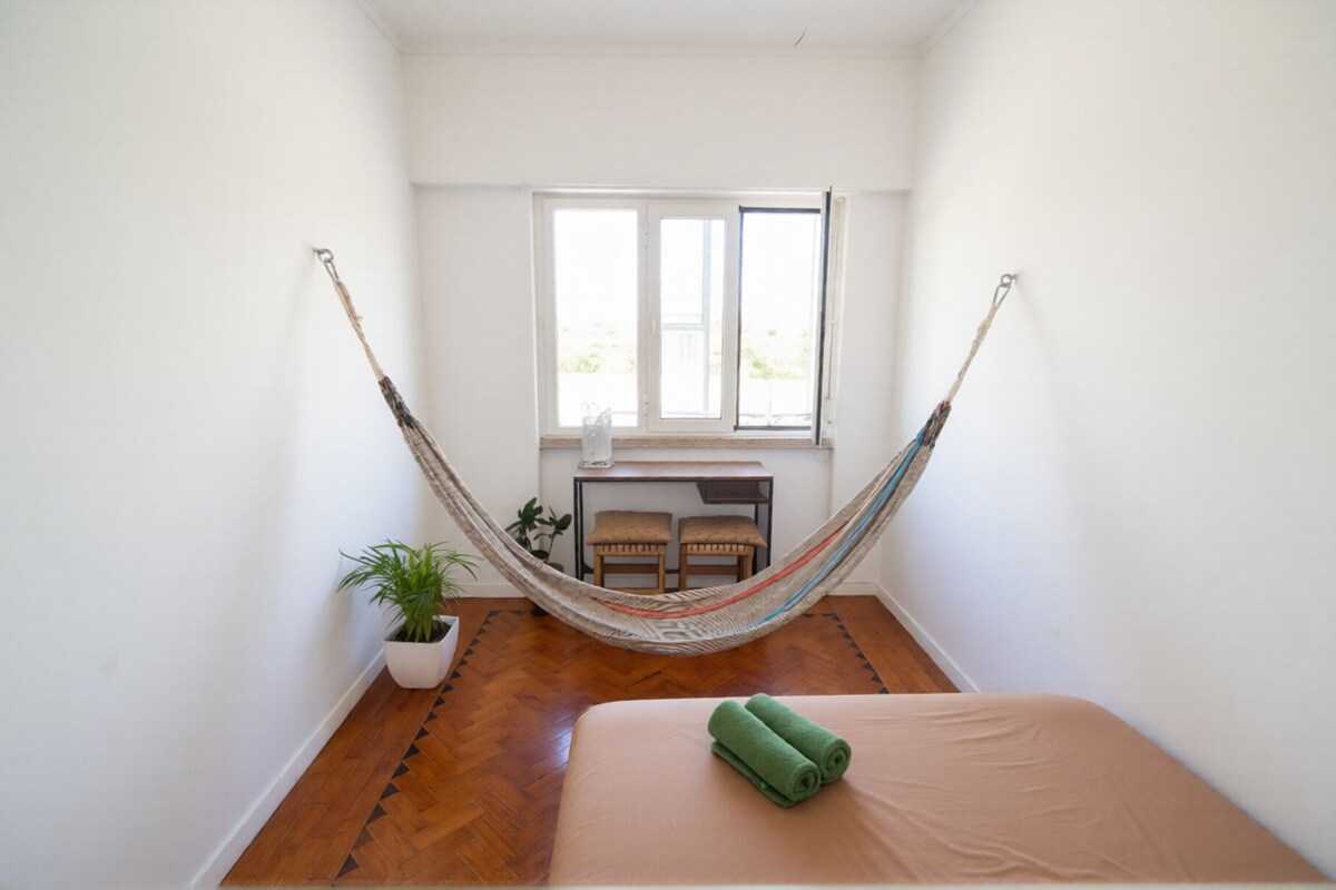 A room with a hammock, a bed, a small table with chairs, and a window.