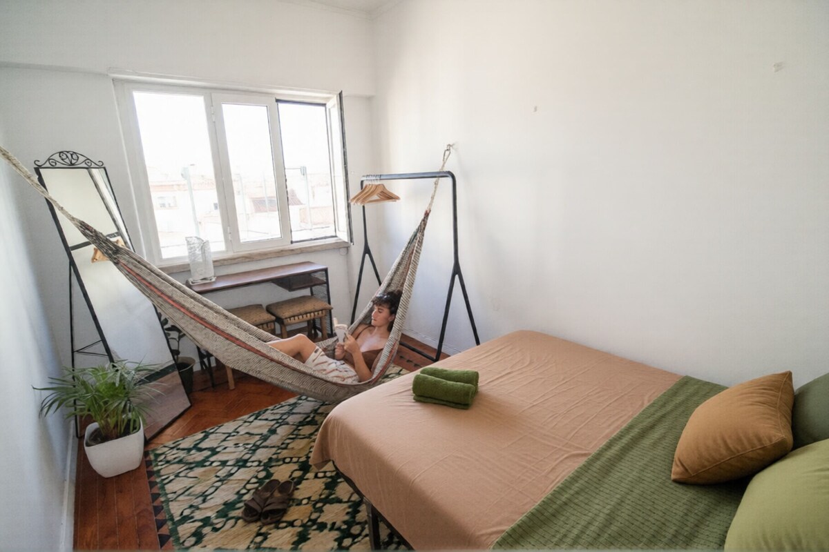 A room with a bed, a hammock, a desk, and a window.