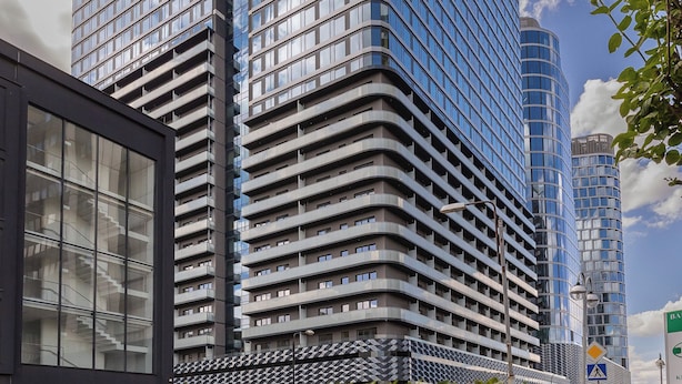 Un immeuble moderne de grande hauteur, doté d’une façade incurvée, entouré de verdure et se dressant sous un ciel partiellement nuageux.