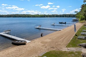 Una playa junto al lago con un muelle para embarcaciones, orilla arenosa y sillas de descanso.
