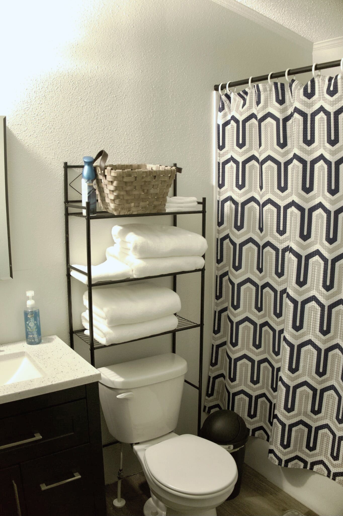 Un baño con inodoro blanco, un estante con toallas dobladas, una cesta y una cortina de ducha estampada.