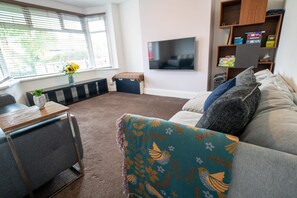 Een woonkamer met een bank, een salontafel, een aan de muur bevestigde televisie en een raam met jaloezieën.