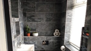 Een badkamer met donkere tegels, een wit wastafelmeubel, een toilet, een klein plankje met planten en een raam met zonwering.