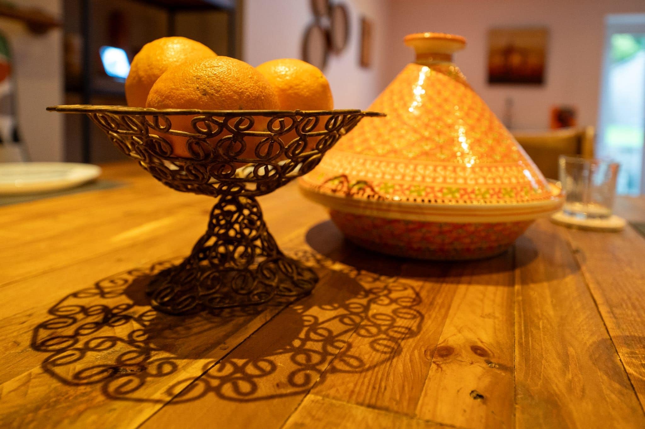A wooden table with a decorative metal bowl holding oranges and a Moroccan tagine.
