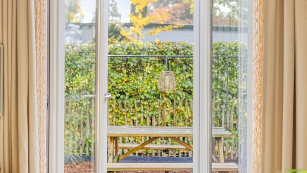 A view through a window with a wooden bench and a hanging basket, with a hedge and trees in the background. - Knusse Tiny house op de Veluwe (Epe)