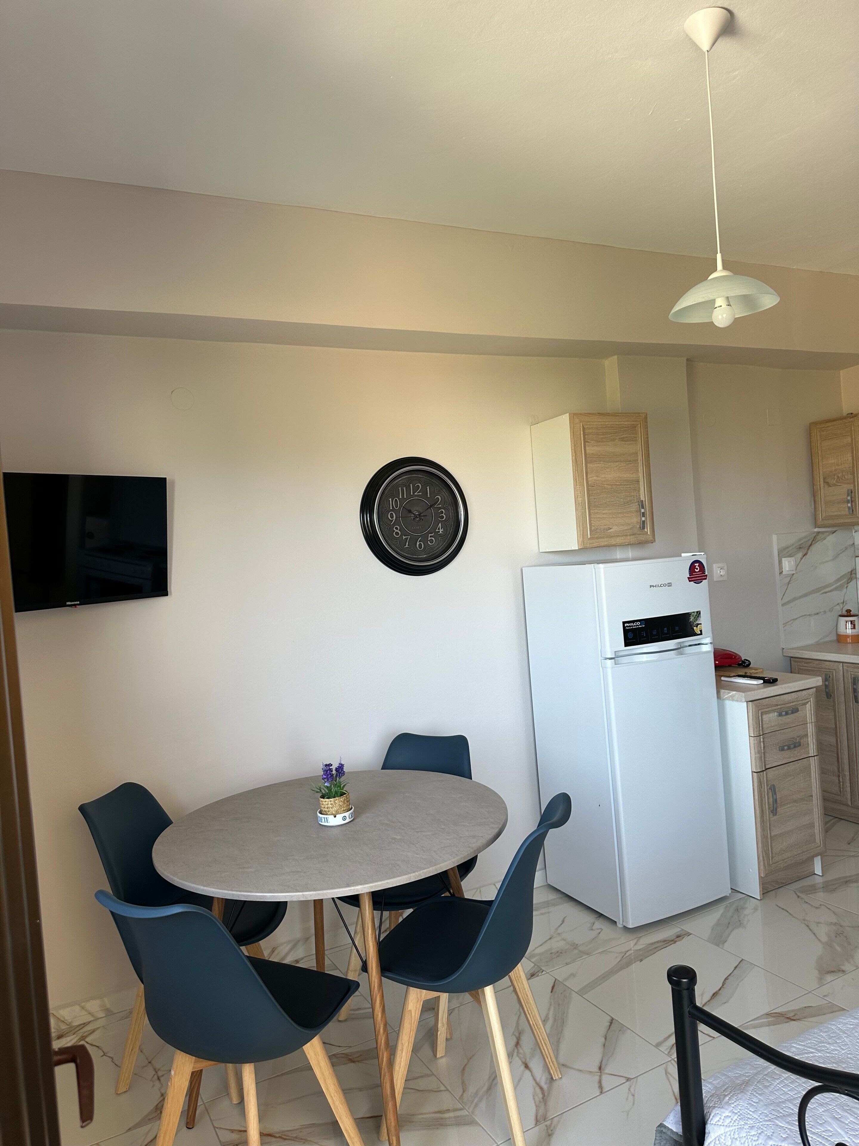 A compact living space with a dining area, a wall-mounted clock, a flat-screen TV, and a white refrigerator.