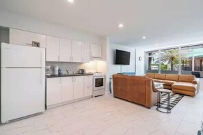 A modern kitchen with white cabinets, a refrigerator, and a dining area with a brown sofa and a TV mounted on the wall. - The Purple Pelican~Beachfront w/Ocean Balcony~#4 (Fort Lauderdale)