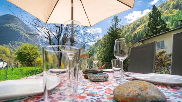 Ein Essplatz draußen mit einem Tisch, der mit einer blumigen Tischdecke, Weingläsern und Geschirr für eine Mahlzeit gedeckt ist.
