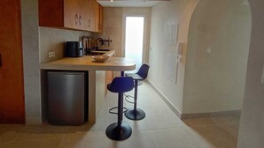 Une cuisine compacte équipée d’un réfrigérateur, d’un évier et d’une cuisinière. On y trouve deux tabourets de bar bleus et une petite table. La cuisine est dotée d’armoires en bois et d’un plan de travail blanc.